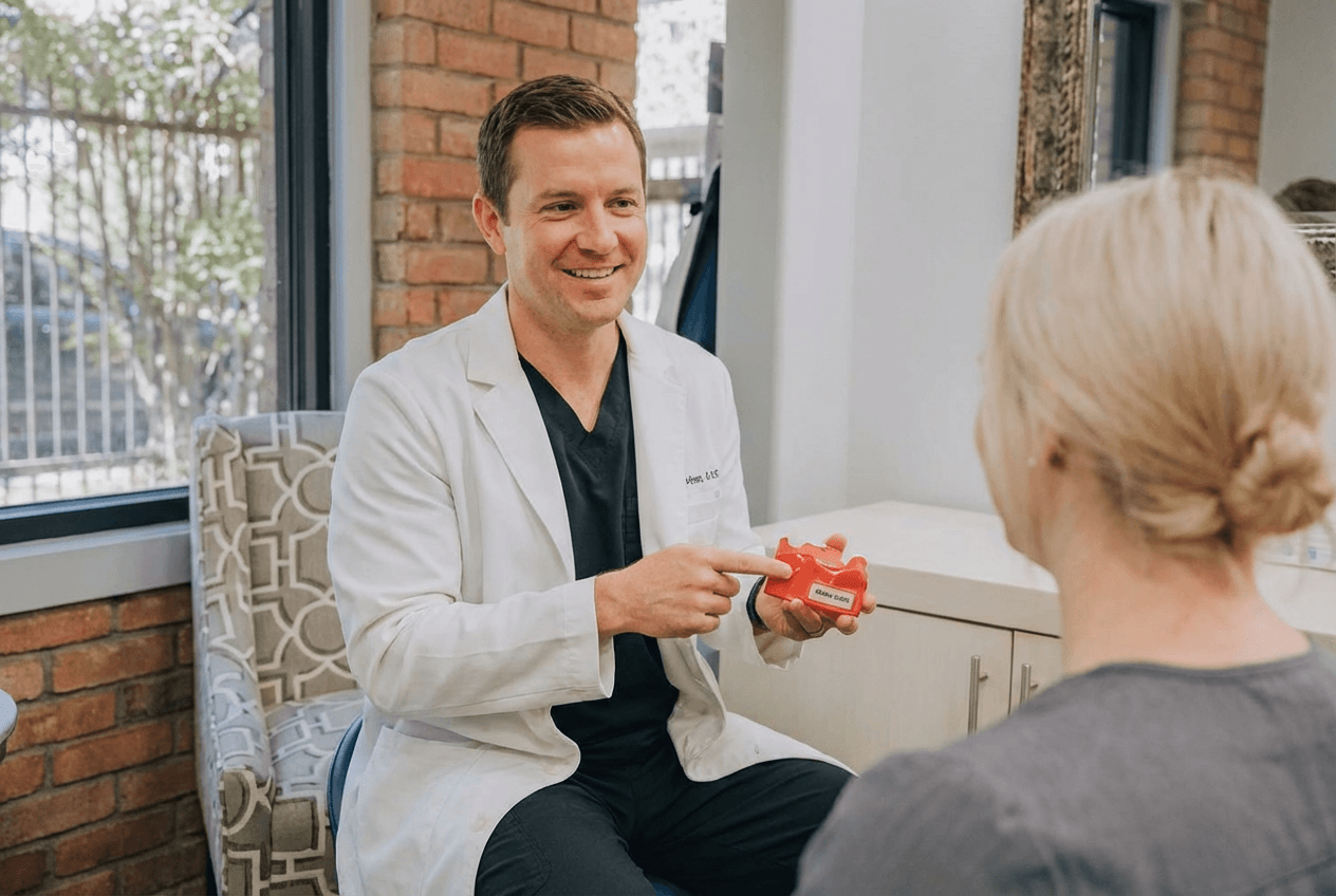A male support specialist in a white lab coat smiling while showing a red dental model of a jaw to a blonde female patient in a clinic setting with a brick wall background.